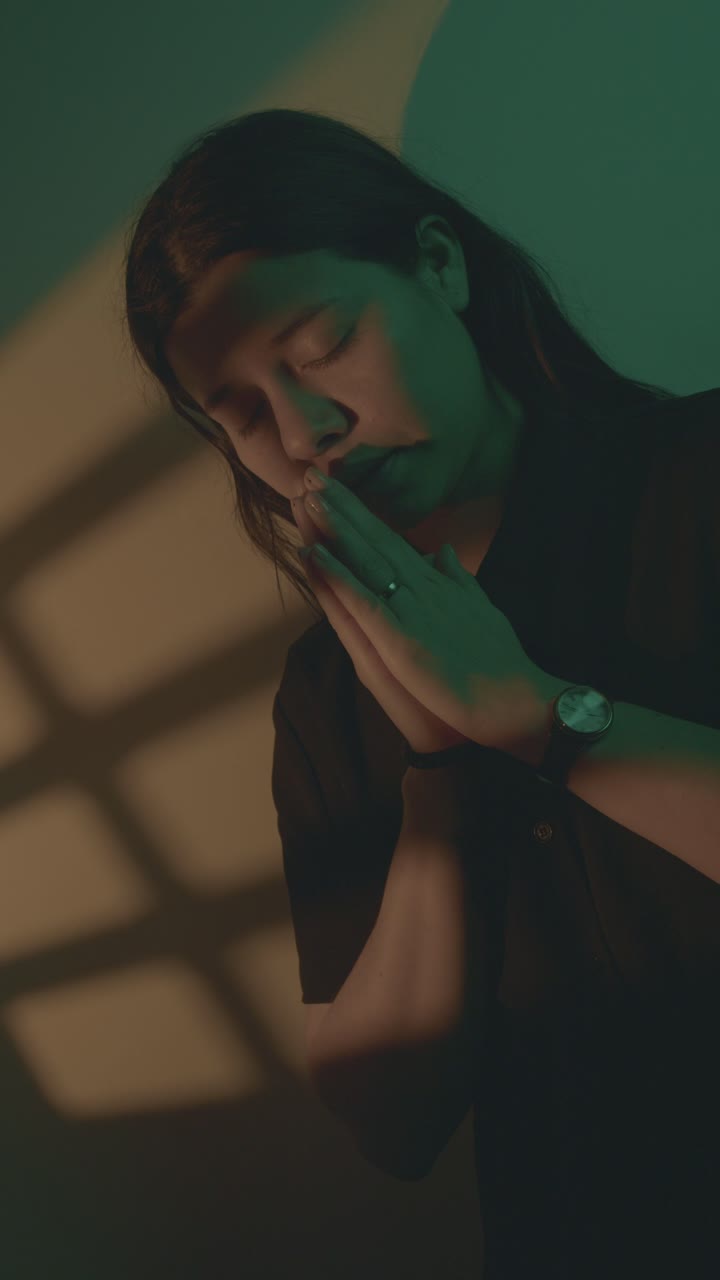 Intimate close-up of a woman praying, hands clasped, in moody, dramatic lighting. Shadows cast by window frame enhance the serene, contemplative mood. Vertical video ideal for social media