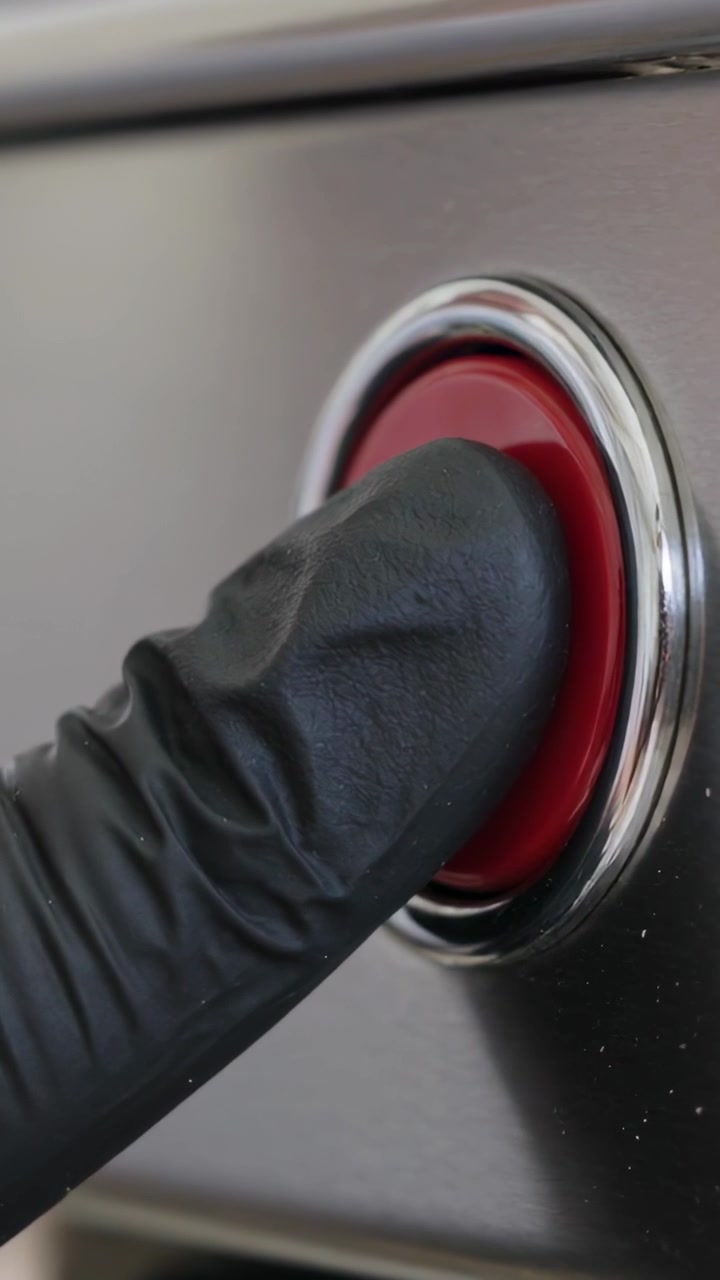 Vidéo verticale : Un doigt ganté appuyant et maintenant un bouton rouge sur un panneau de commande métallique.