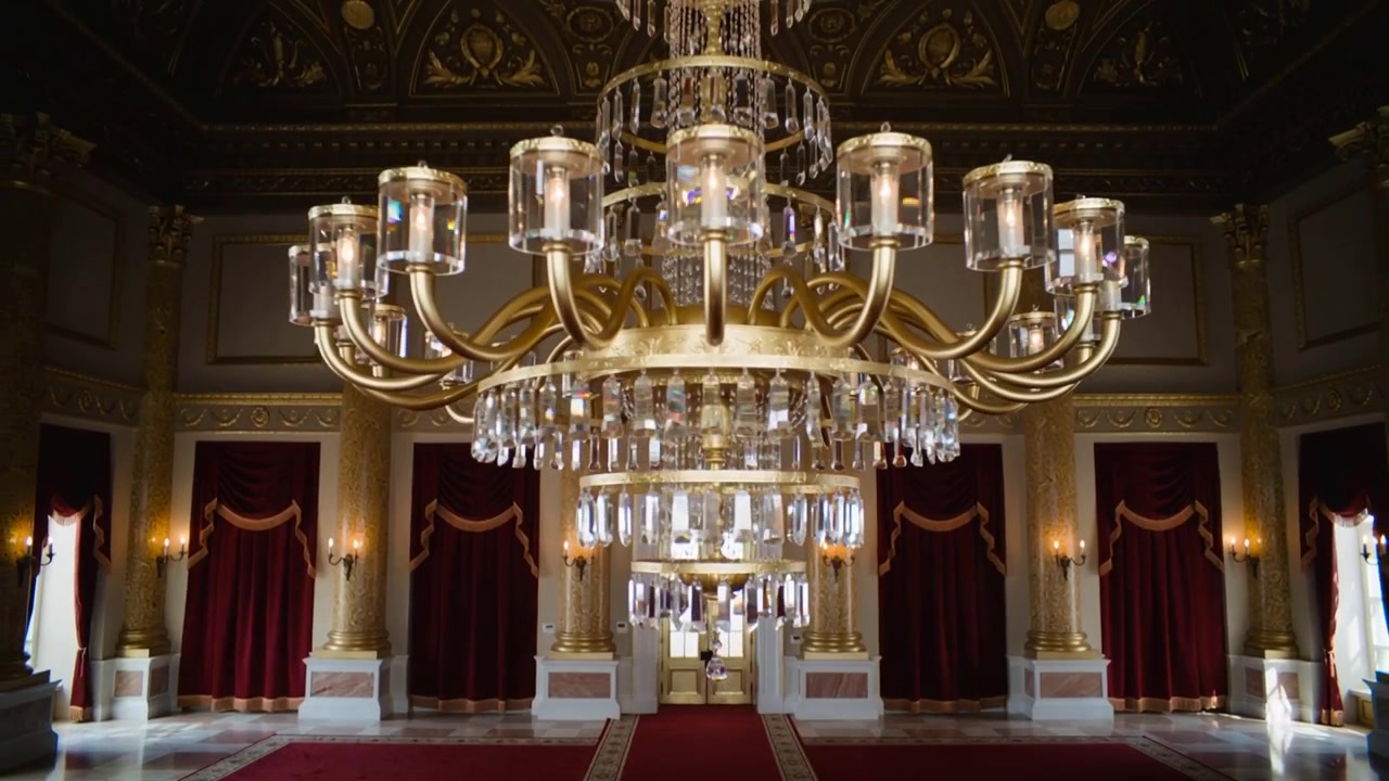 Telecamera che si allontana rivelando un lampadario di cristallo a più livelli nella grande sala di un palazzo, con una passatoia rossa.