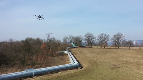 Quadcopter-Drone-Aircraft-Flying-Above-Pipelines-and-Meadow-on-Sunny-Day,-Aerial-Tracking-View