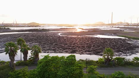 drone push in over road and tropical trees to asphalt runoff lake with oil refinery behind snaking river of water