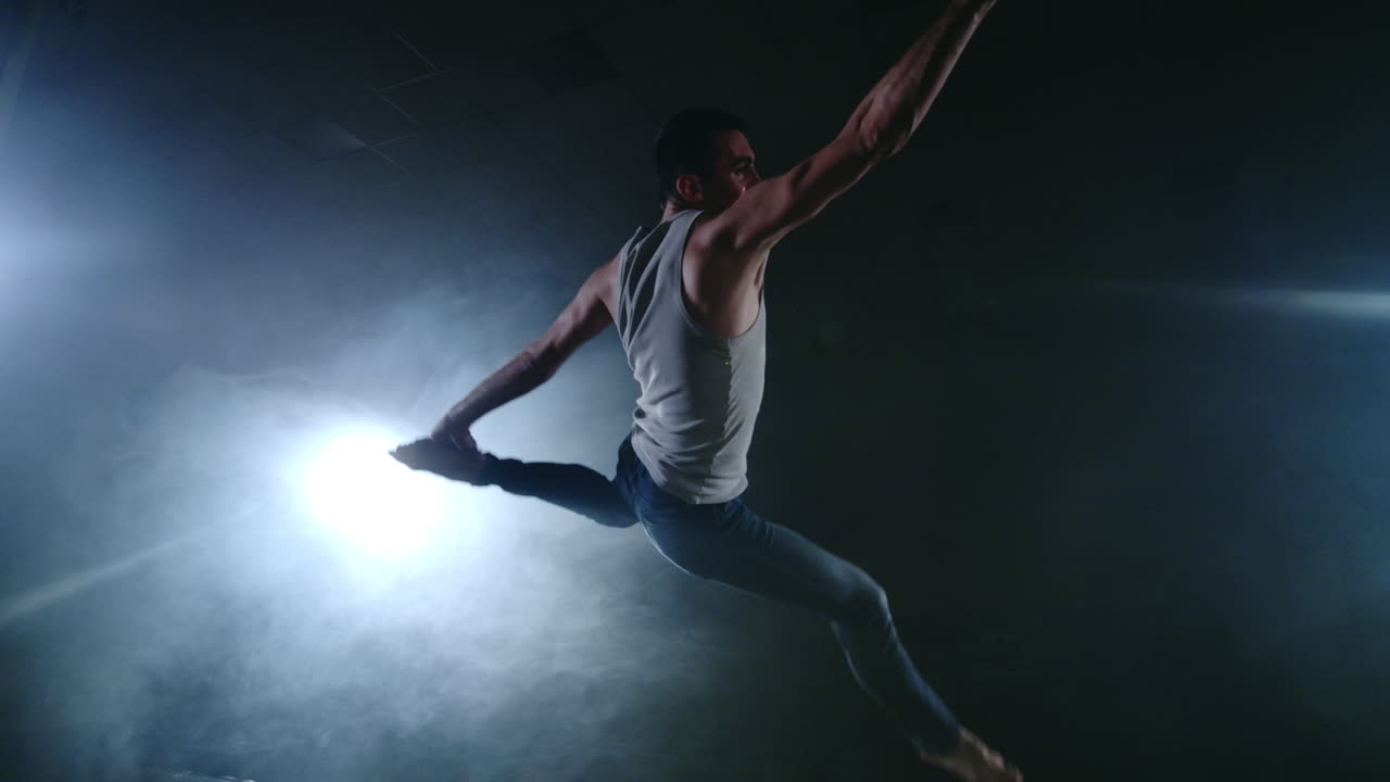 A modern ballet a man performs jumps and spins in the light of spotlights and smoke on a dark background. Acrobatic choreography rehearsal of the script of modern ballet.