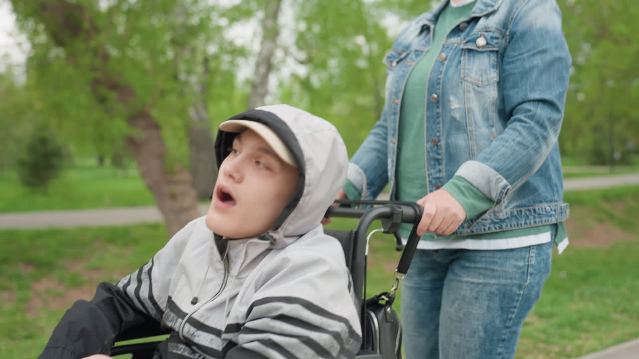 Serene Walk Through Lush Green Park Environment, Relaxed Woman Helps Teen Navigate Leafy Park Pathway Peacefully, Calm Outdoor Scene With Woman Pushing Teen In Wheelchair Along Vibrant Park Trail