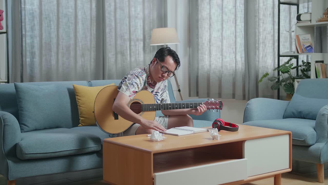 Asiatisk kompositör med gitarr skrynklar ihop papper och kastar bort det, får sedan huvudvärk medan han komponerar musik hemma