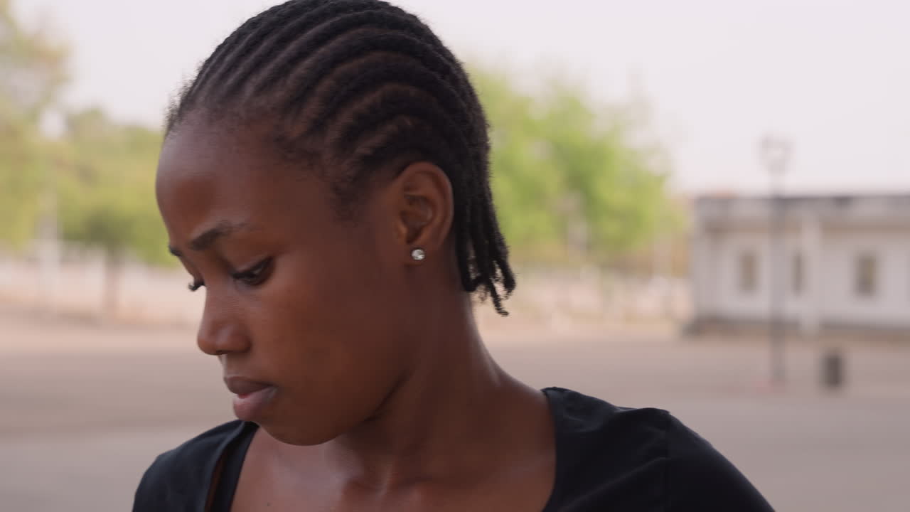 Giovane donna all'aperto, in una strada vuota, con capelli intrecciati e orecchino a perno. Guarda di lato con espressione pensierosa sotto la calda luce del sole, con edifici distanti e alberi sfocati sullo sfondo e una leggera sudorazione sulla pelle.