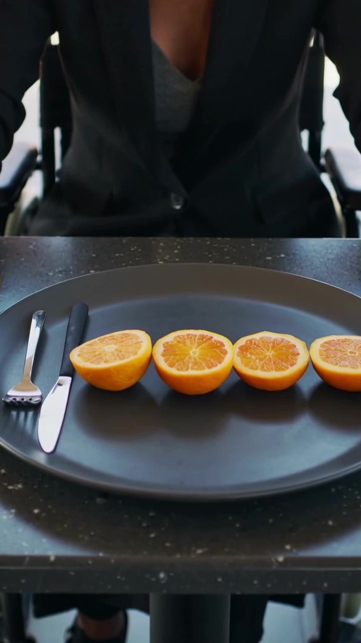 Video vertical: Trayendo un plato, mujer en silla de ruedas con blazer en la mesa alineando cuatro mitades de naranja