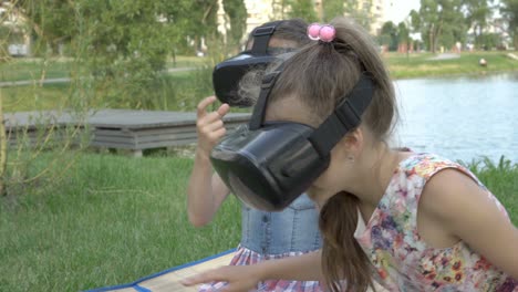 primer plano, dos chicas jóvenes jugando juegos virtuales con gafas negras de realidad virtual, sentadas en el patio trasero junto al río en una noche de verano. el concepto de unas vacaciones en cuarentena.