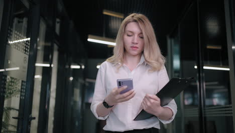 Smiling-businesswoman-looking-on-cellphone-indoors.-Surprised-business-woman-reading-message-on-mobile-phone-in-office-corridor.-woman-looking-smartphone-screen-in-business-center.-walking-office