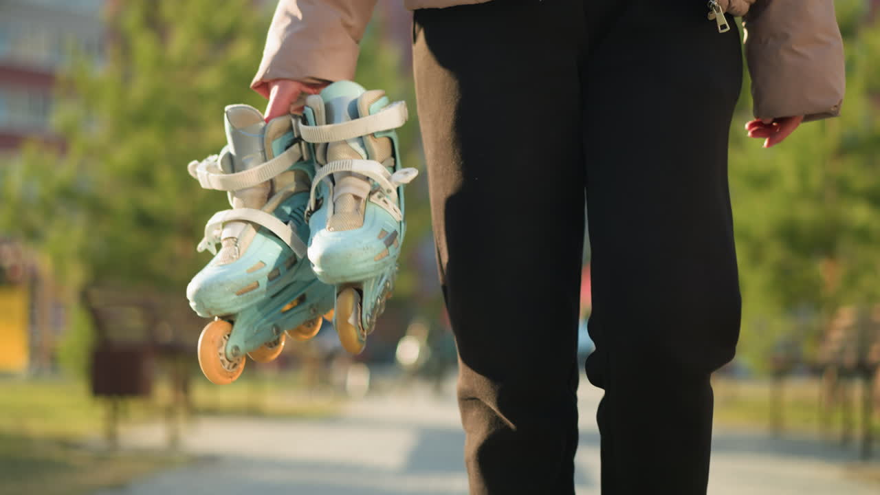 Front shot of a person holding a pair of rollerblades, wearing black trousers and a peach jacket, walking through a sunlit park with a blurred background of trees. The person's face is not visible