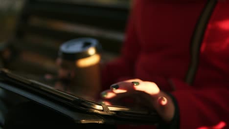 mujer de rojo sosteniendo café en una mano y escribiendo en una tableta mientras se sienta en un banco en el parque durante el clima otoñal