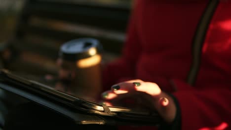 woman in red holding coffee in one hand and typing on tablet while sitting on bench in park during autumn weather