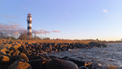 Leuchtturm-Bei-Sonnenuntergang-Und-Wellen,-Die-Auf-Felsen-Schlagen