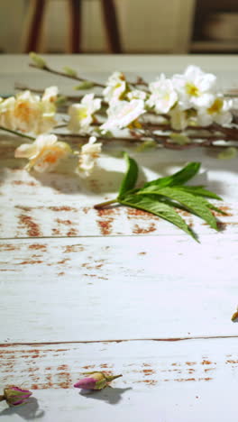 a female hand lifts a bubble-shaped glass of pink iced fruit tea—freshly stirred with syrup—from a whitewashed wooden table sprinkled with dried blossoms, bathed in warm morning sunlight.