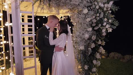 recién casados, novio, novia abrazando, abrazando, besando, ceremonia de la noche de la boda, arco con flores