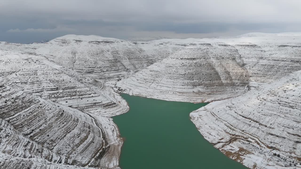 Kış mevsiminde Lübnan'daki Chabrouh Barajı'nın turkuaz suyu, karla kaplı kayalık seti ve huzurlu atmosferi gösteren sinematik havadan görünümü.