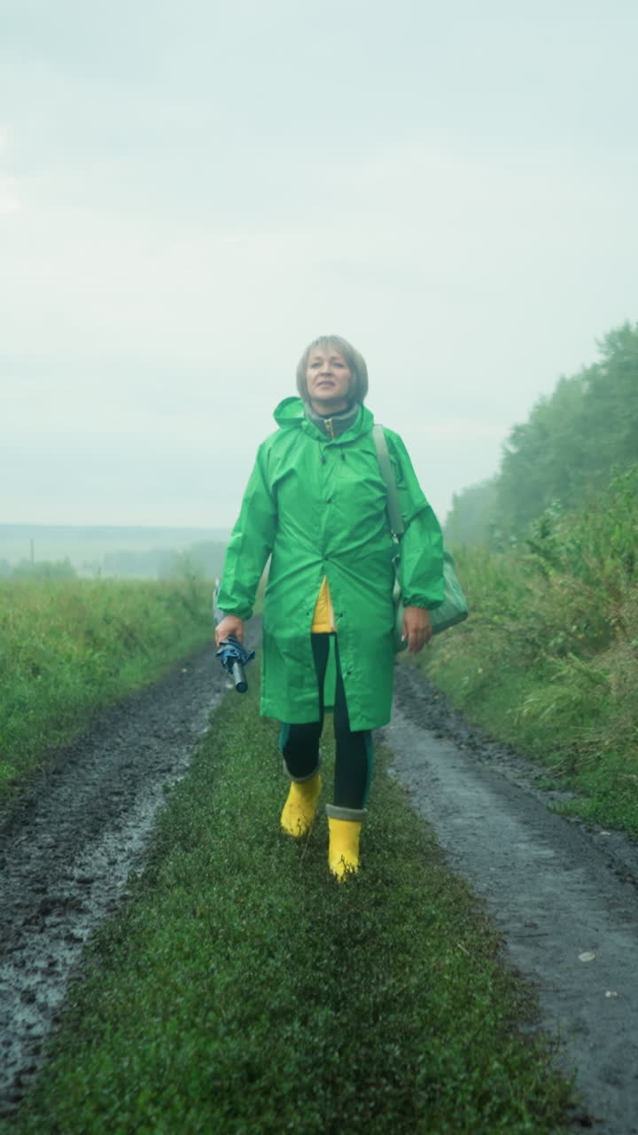 Woman in green raincoat carrying mint-colored bag and umbrella, wearing yellow rain boots, walking along a muddy path surrounded by greenery and trees under a misty sky