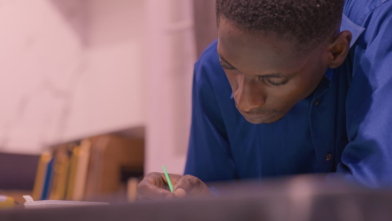 Black student leaning over desk writing with green pen, blue shirt and books in background, closeup study session showing note taking, sketching diagrams and concentrated reading in quiet indoor
