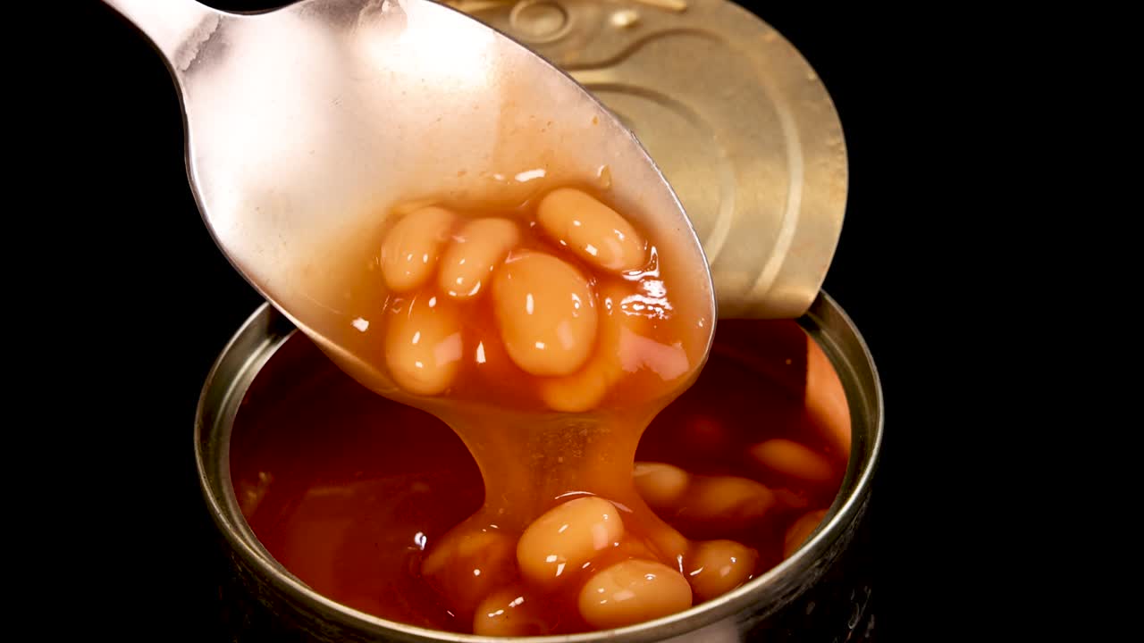 A metal spoon lifts baked beans in tomato sauce from an open can under bright, direct lighting with a black background and minimal camera movement