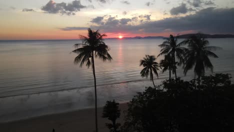 red colored sunset on the horizon above the silhouettes of the mountains between the high green palmtrees while people are walking on the sandy beach near krabi