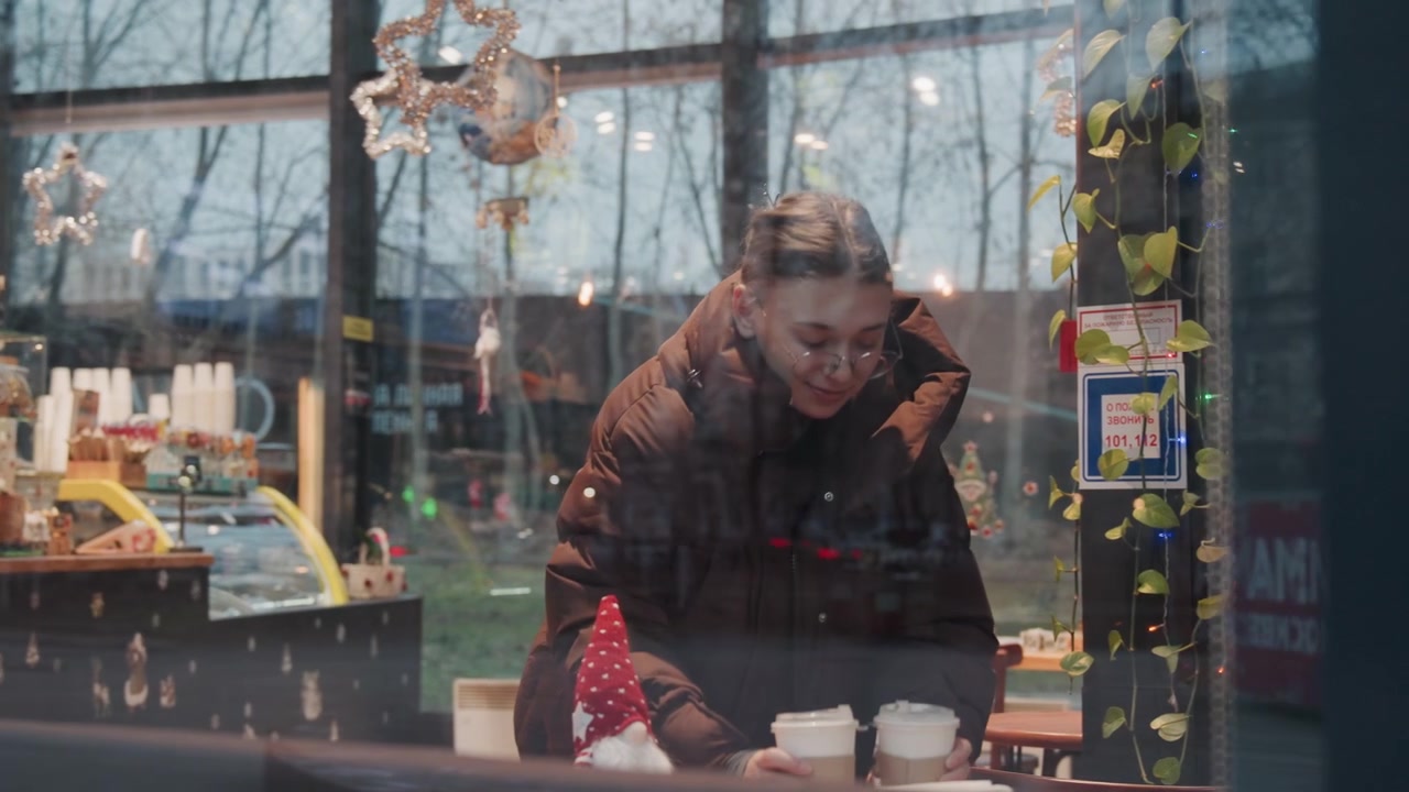 Regennasses Fenster, weiße Frau trägt zwei Kaffee zum Mitnehmen in ein Café, trägt einen dicken Parka, Spiegelungen von Straßenlaternen auf dem Glas, Pendlerin balanciert Tassen, während sie sich darauf vorbereitet, den Mantel auszuziehen, geschäftige urbane Stimmung