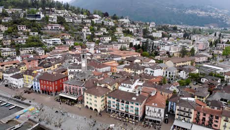 Aerial-flyover-with-a-turn-from-the-shores-of-Lago-Maggiore-over-the-promenade-of-Ascona-in-Ticino,-Switzerland-over-the-rooftops-around-the-church-tower-revealing-the-lake-and-mountains