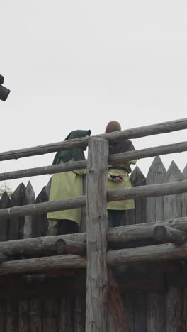 couple of people stands guard at edge of medieval settlement. sentinels watch area outside village looking over high wooden fence low angle shot backside view