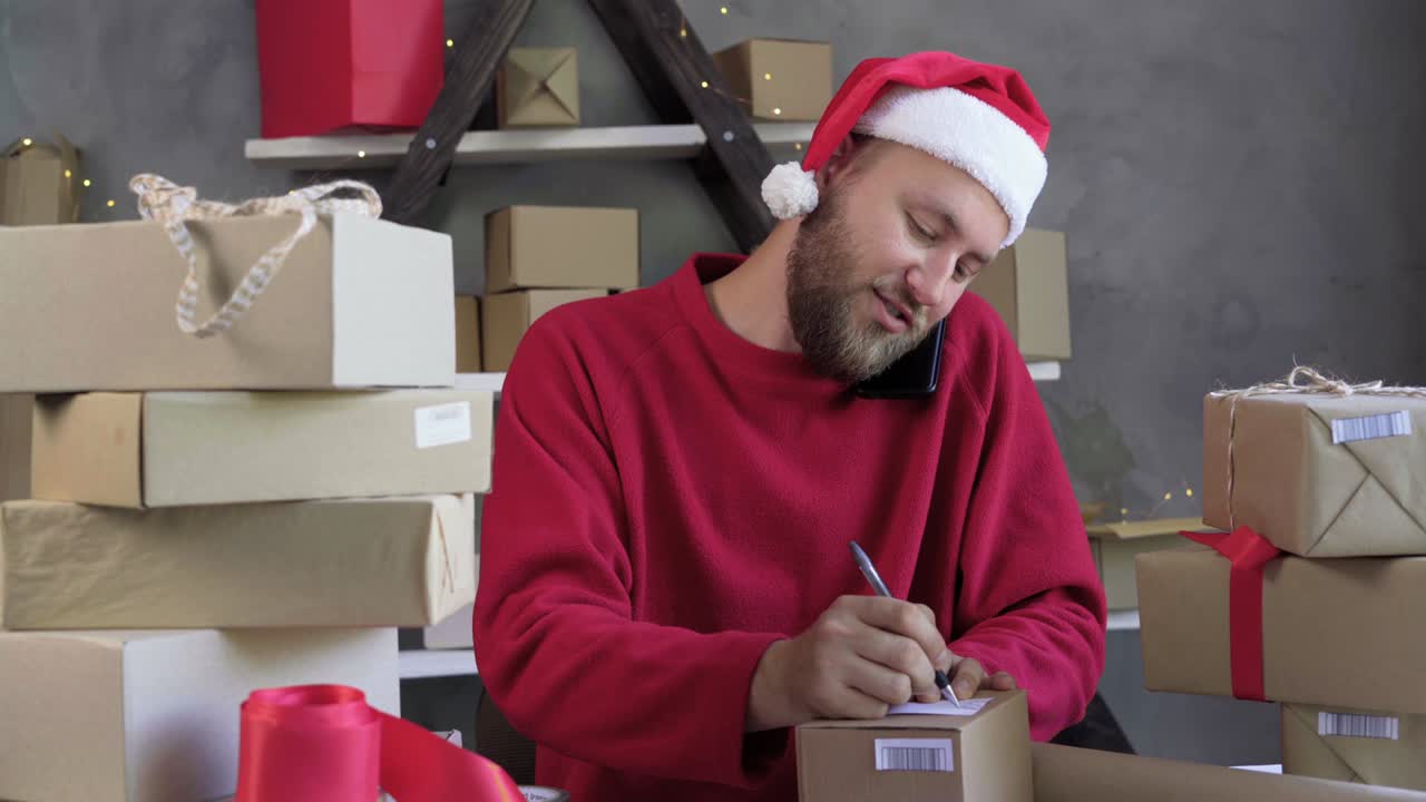 male entrepreneur business owner working at home on christmas eve wearing a red santa claus hat and sweater received a phone call from a customer and writes an address for delivery.