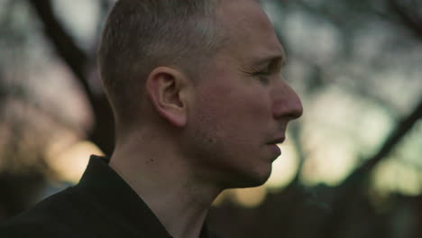 a close-up side view of a man in a green jacket exhaling smoke through his nose and mouth at dusk. the blurred background with trees and soft light