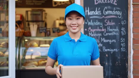 a young woman in a blue shirt and cap carries a box of fresh pastries outside a local bakery. the shop displays delicious treats, inviting hungry customers to stop by.