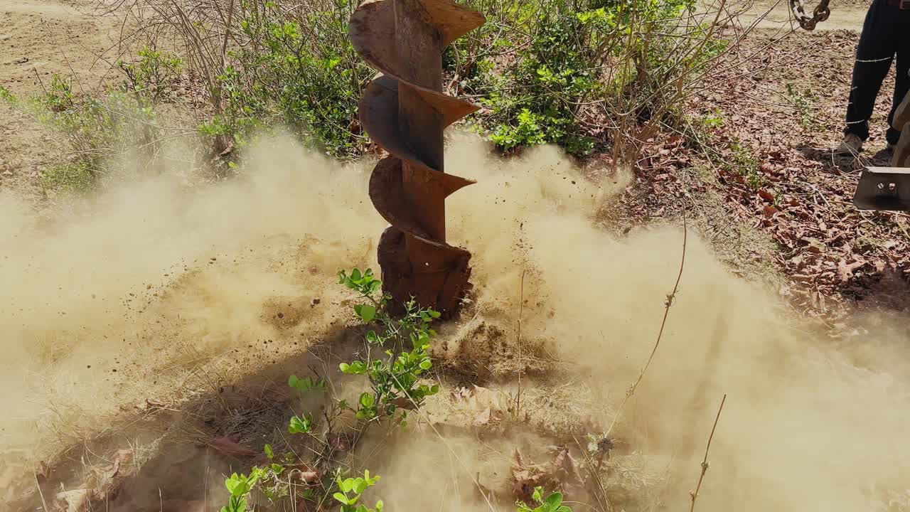 Primo piano di una trivella meccanica che fora terreno asciutto su terreni agricoli, creando un foro per l'installazione di pali di cemento e recinzioni di filo spinato, con la polvere che si alza alla luce del sole