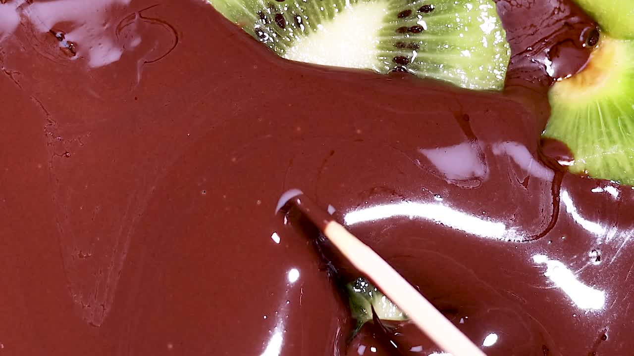 Close-up of kiwi slices being enveloped by smooth, melted chocolate with a stick for dipping.