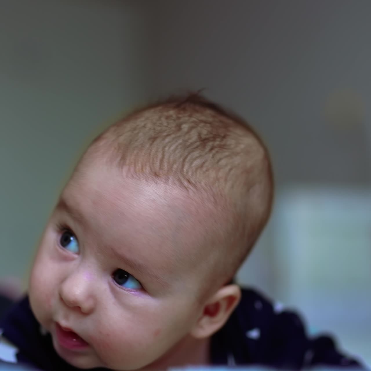 Cute baby lies on bed on his stomach. Beautiful infant child looks up with curiosity. Close up. Blurred backdrop