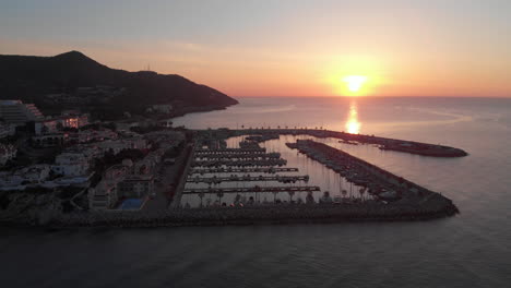 Sunrise-drone-shot-with-fishing-port-surrounded-by-serene-waves-near-coastline-city-with-mountain-range-skyline