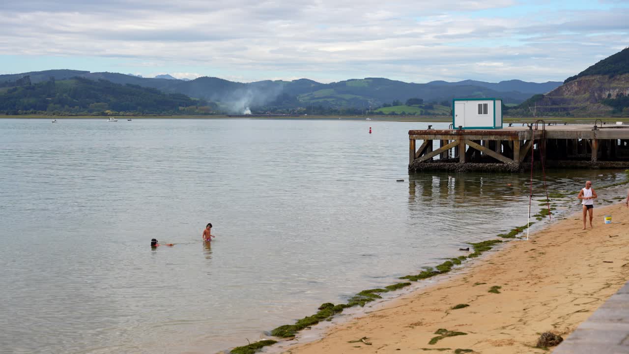 Kids playing in the shallow waters near a quiet pier with scenic mountains in background