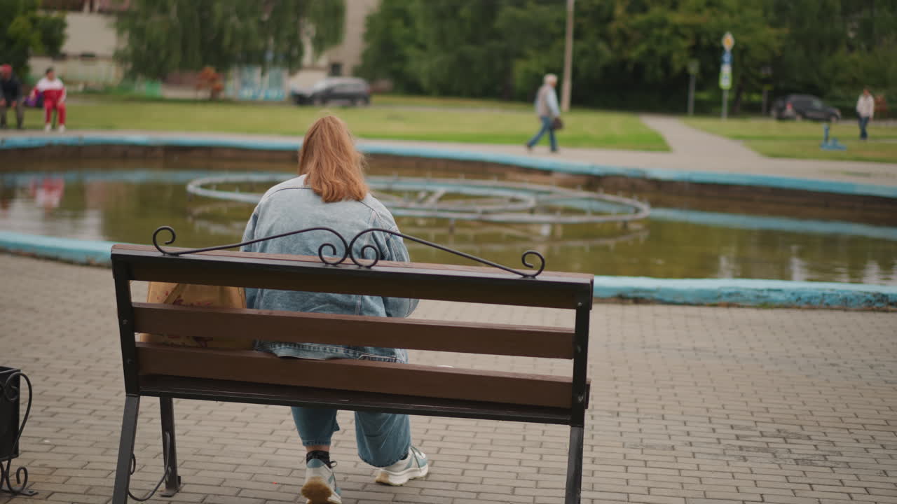 Kavkazská žena sedící na lavičce u fontány, džínová bunda, zrzavé vlasy, ležérní džíny, taška vedle lavičky, lidé procházející v pozadí, dlážděné náměstí, jemné denní světlo, klidná zamyšlená nálada, městský park.