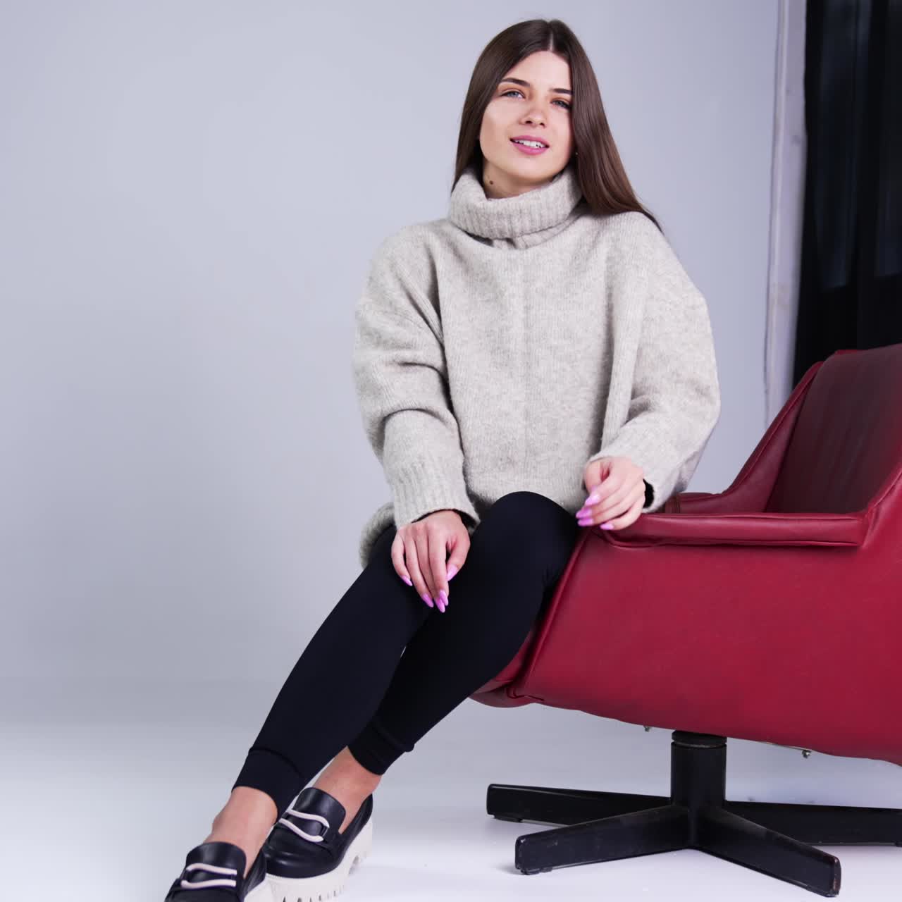 Young beautiful long-haired brunette wearing warm sweater, tight pants and stylish shoes sitting in red armchair indoors. Model posing in the studio