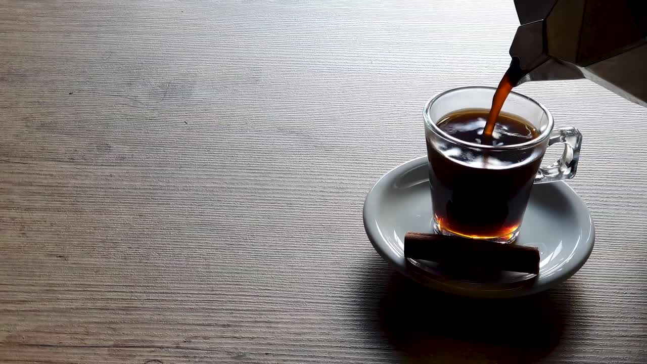 Pouring coffee in empty transparent cup, supported by white saucer on wooden table