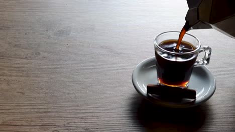 Pouring-coffee-in-empty-transparent-cup,-supported-by-white-saucer-on-wooden-table