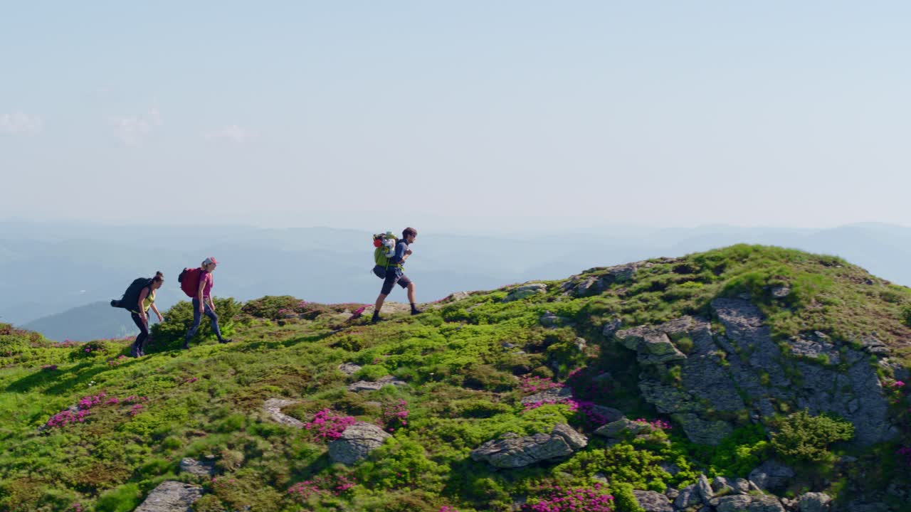 Aerial shot of three backpackers reaching a mountain summit viewpoint. Conveys adventure, achievement, freedom, and extreme sports. Filmed on a clear, sunny day
