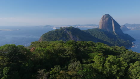 Aerial-footage-flying-over-the-trees-on-a-mountain-top,-revealing-the-cable-lift-of-Sugarloaf-mountain-in-the-background