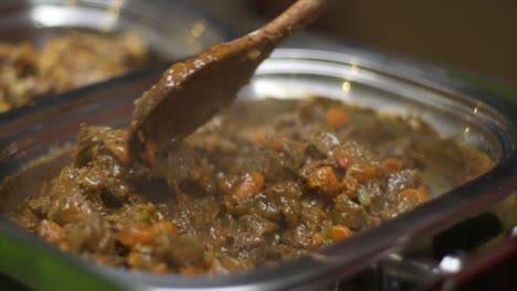 Meat-curry-in-stainless-steel-warming-tray-being-mixed-and-stirred-using-wooden-spoon,-filmed-as-close-up-in-slow-motion