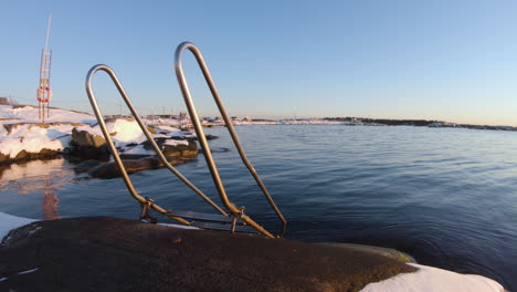 4k-Close-up-of-a-metal-staircase-by-snowy-rocks-and-the-very-cold-ocean-water-during-a-winter-day-at-Gothenburg,-Sweden