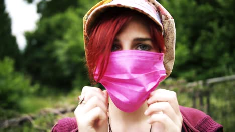 young red haired girl with colorful cap puts on a medical pink face mask with green environment in background in slow motion