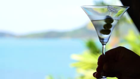 a hand holds a martini glass with olives against a vibrant ocean backdrop, capturing a serene and refreshing moment