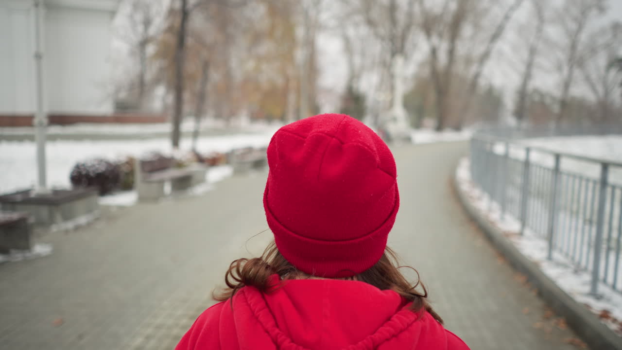 Close-up of woman jogging along snowy park pathway in red hoodie and black leggings, surrounded by serene winter scenery with benches, bushes, iron railing, lamp posts, and distant cross monument