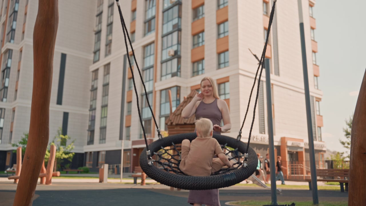 Una donna spinge delicatamente un bambino su un'altalena a pneumatico in un parco giochi all'aperto, con edifici residenziali sullo sfondo. La scena cattura la loro interazione e il loro movimento mentre si godono il tempo insieme all'aperto.