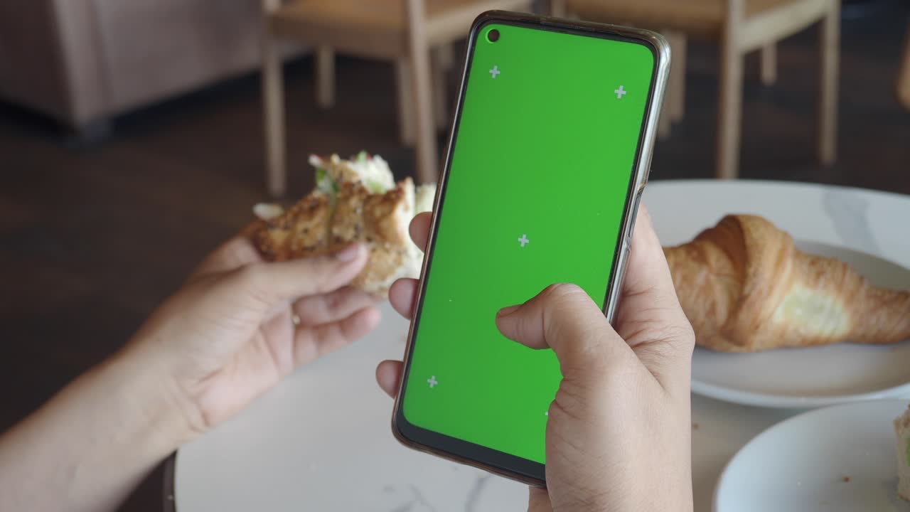 mujer usando un teléfono con pantalla verde mientras come sándwich y croissant en una cafetería