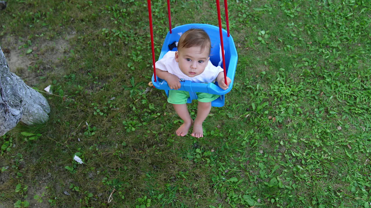 Thoughtful little baby sitting in a blue swing and swaying. Summer in the village concept. View from above.