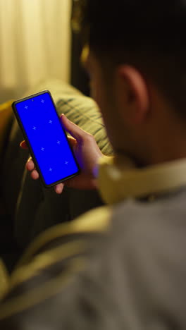 vertical video close up over the shoulder shot of man spending evening at home sitting on sofa looking at blue screen mobile phone