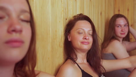 close up of women enjoying steamy sauna session as one woman fans herself while others smile and watch with relaxed expressions, wooden interior glowing warmly around their calm and peaceful presence
